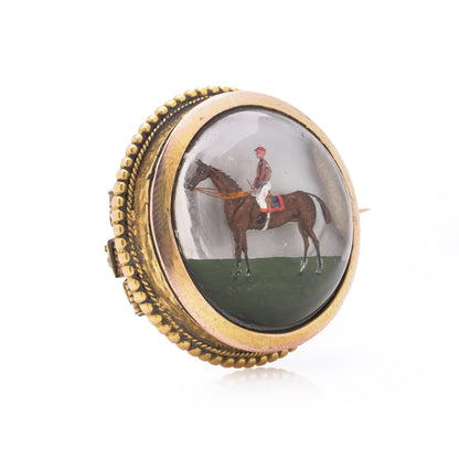 A round crystal brooch in yellow gold featuring a jockey sitting on a horse's back, on a white background
