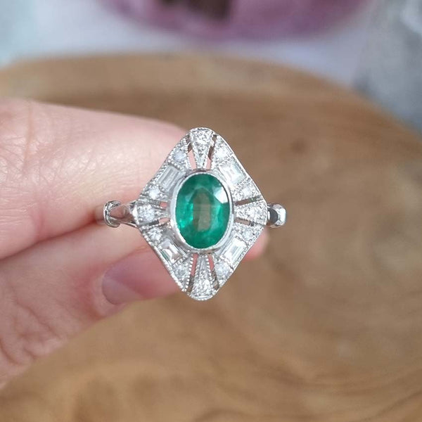 An emerald and diamond navette shaped cluster ring in white metal, held by a hand