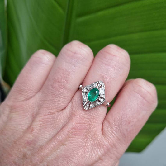 An emerald and diamond navette shaped cluster ring, modelled on a middle finger, with a leaf in the background