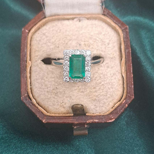 An emerald and diamond rectangular cluster ring in white gold, in an antique ring box, against a green background