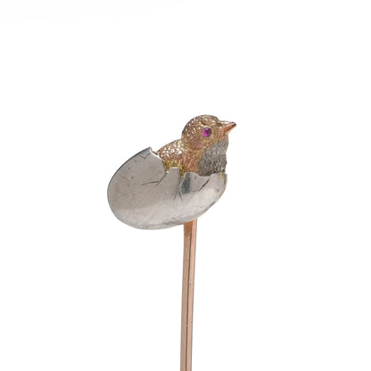 A stick pin depicting a chick hatching from an egg, in gold and silver