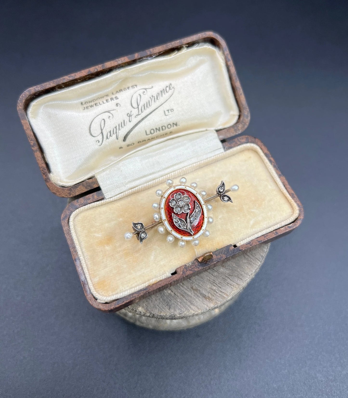 A yellow gold bar brooch with a red enamel centre surrounded by pearls, with a diamond flower motif and diamond leaves at either end, in a box