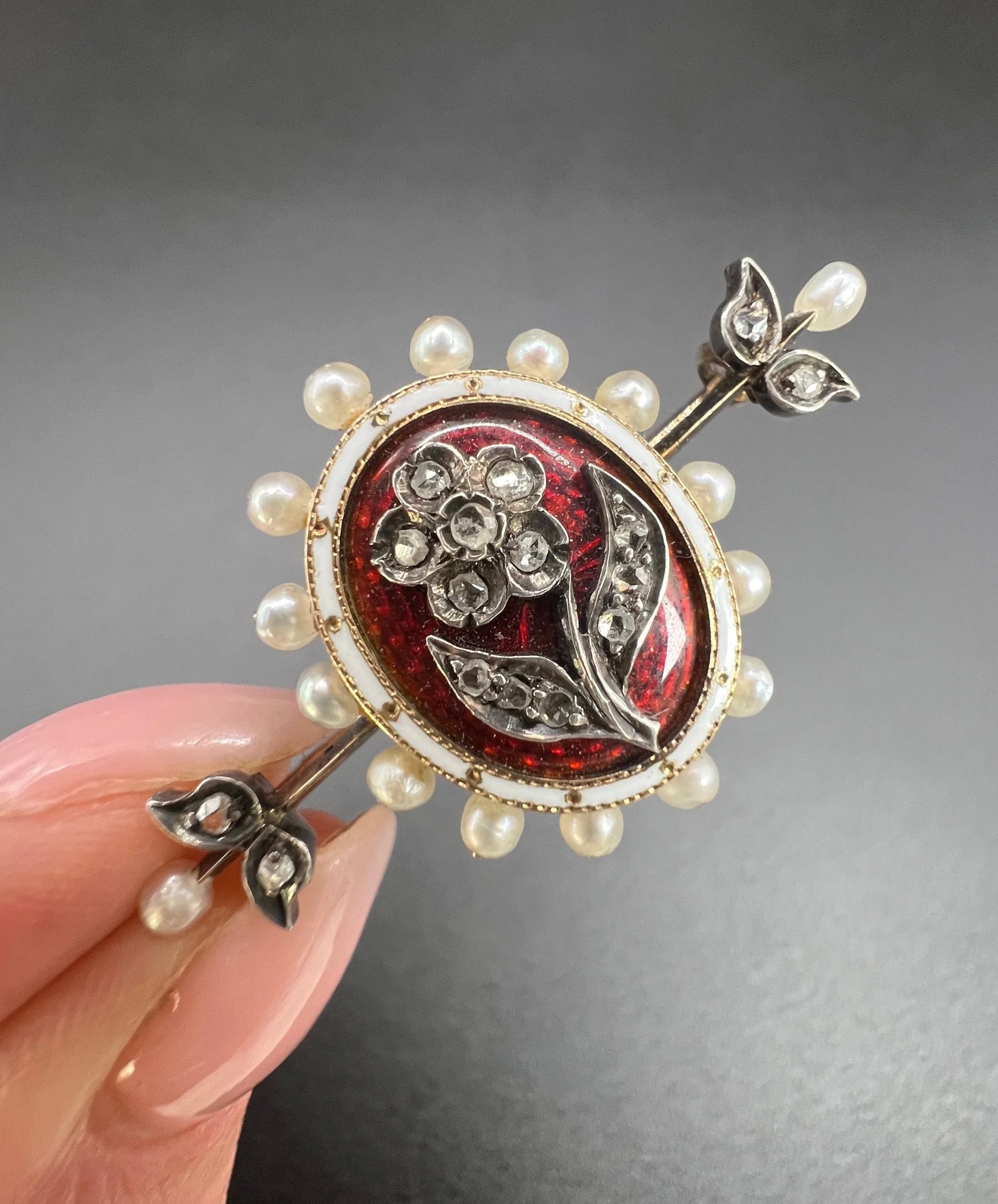 A yellow gold bar brooch with a red enamel centre surrounded by pearls, with a diamond flower motif and diamond leaves at either end, held in a hand