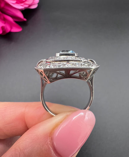 An octagonal shaped cluster ring with an aquamarine centre surrounded by two rows of diamonds, in platinum, held in a hand