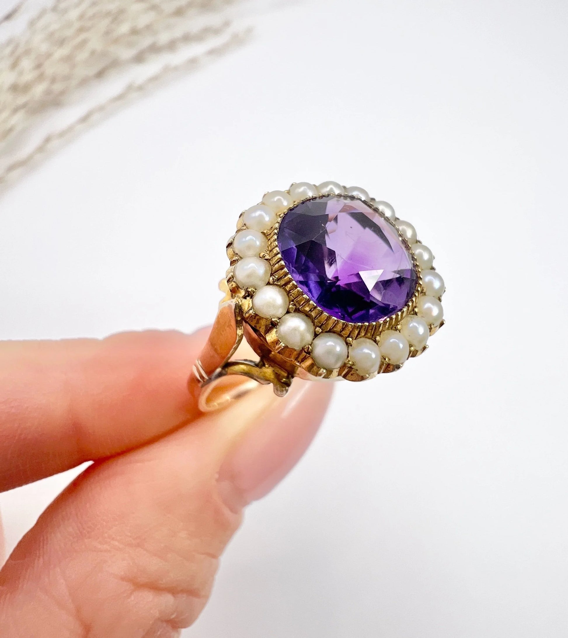 A cushion shaped amethyst cluster ring with a pearl border, in yellow gold, held in a hand