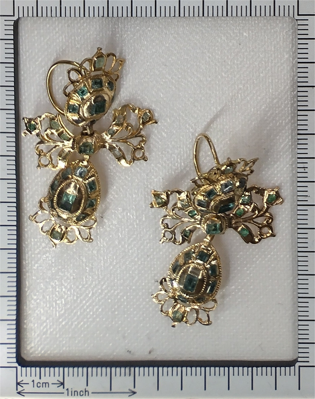 A pair of intricate yellow gold pendant drop earrings set with emeralds, next to a ruler