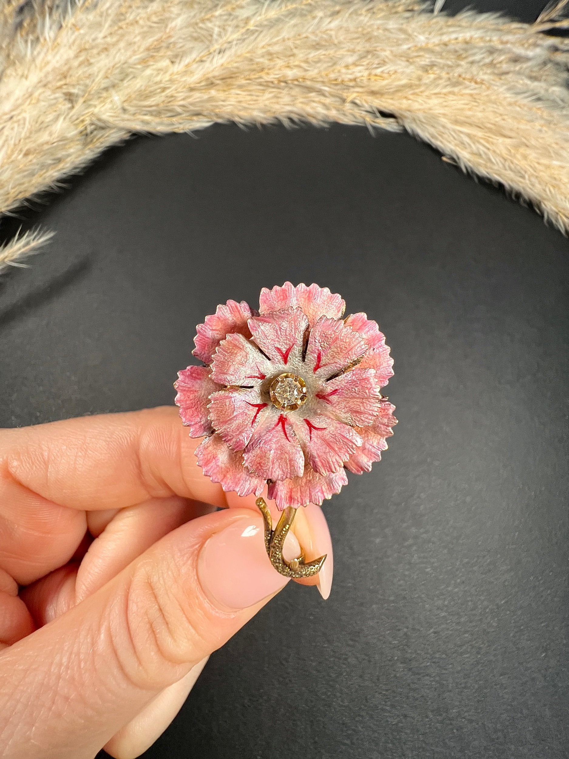 A pink enamelled flower brooch with a diamond centre, in yellow gold, held in a hand