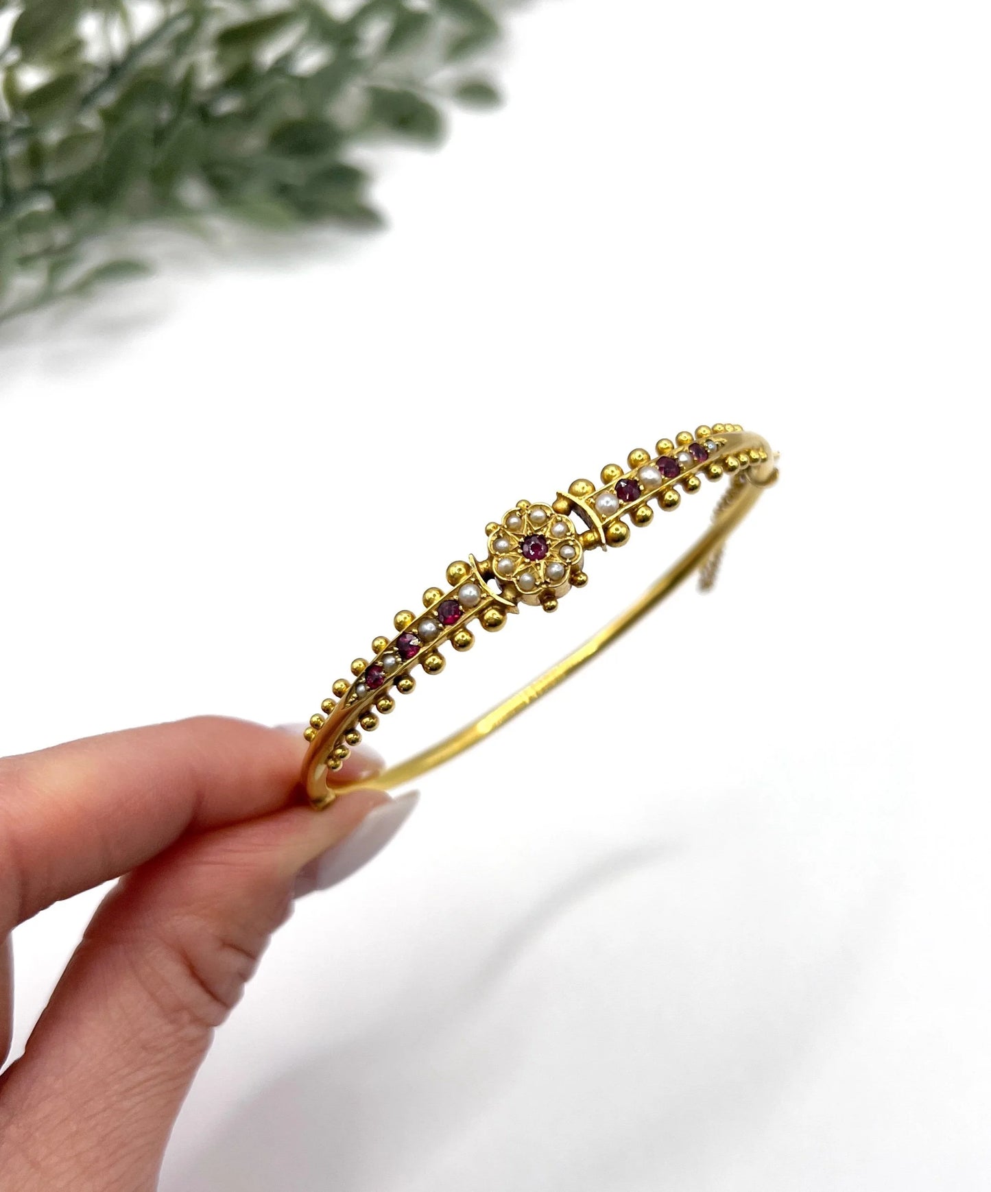 A yellow gold bangle bracelet set with rubies and seed pearls, held in a hand