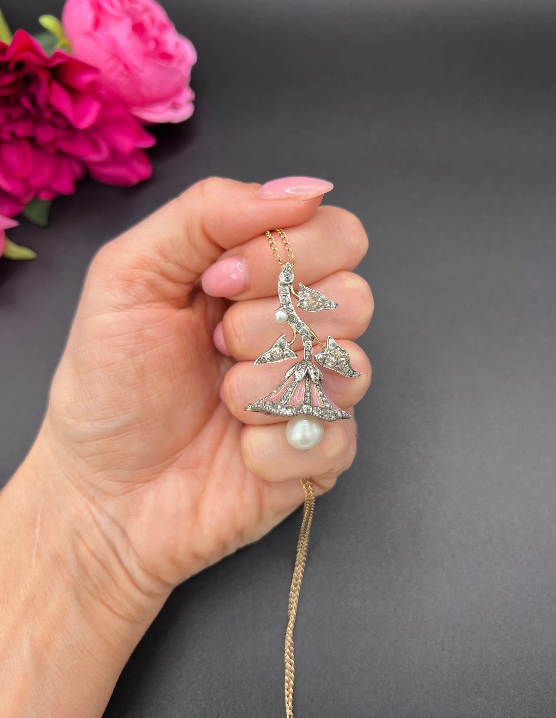 A pink enamel, diamond and pearl flower pendant in gold and silver, with a chain held in a hand