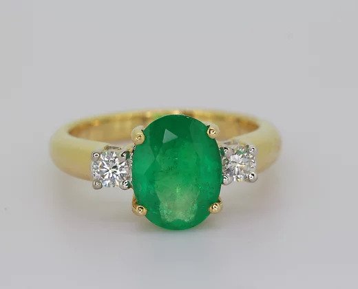 Oval Emerald and Diamond Three Stone Ring; pretty trilogy ring, central oval-cut emerald flanked by brilliant-cut diamonds. Crafted from 18ct yellow gold.