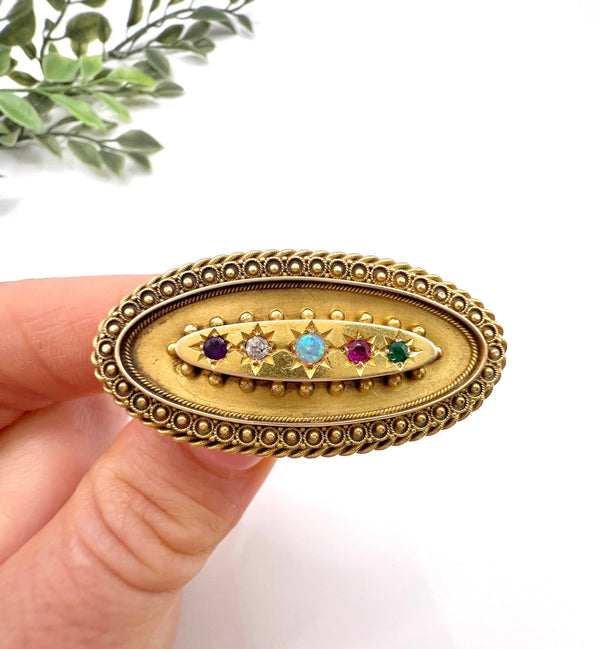 An oval yellow gold multi-gemstone acrostic brooch, held in a hand