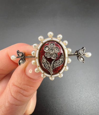 A yellow gold bar brooch with a red enamel centre surrounded by pearls, with a diamond flower motif and diamond leaves at either end