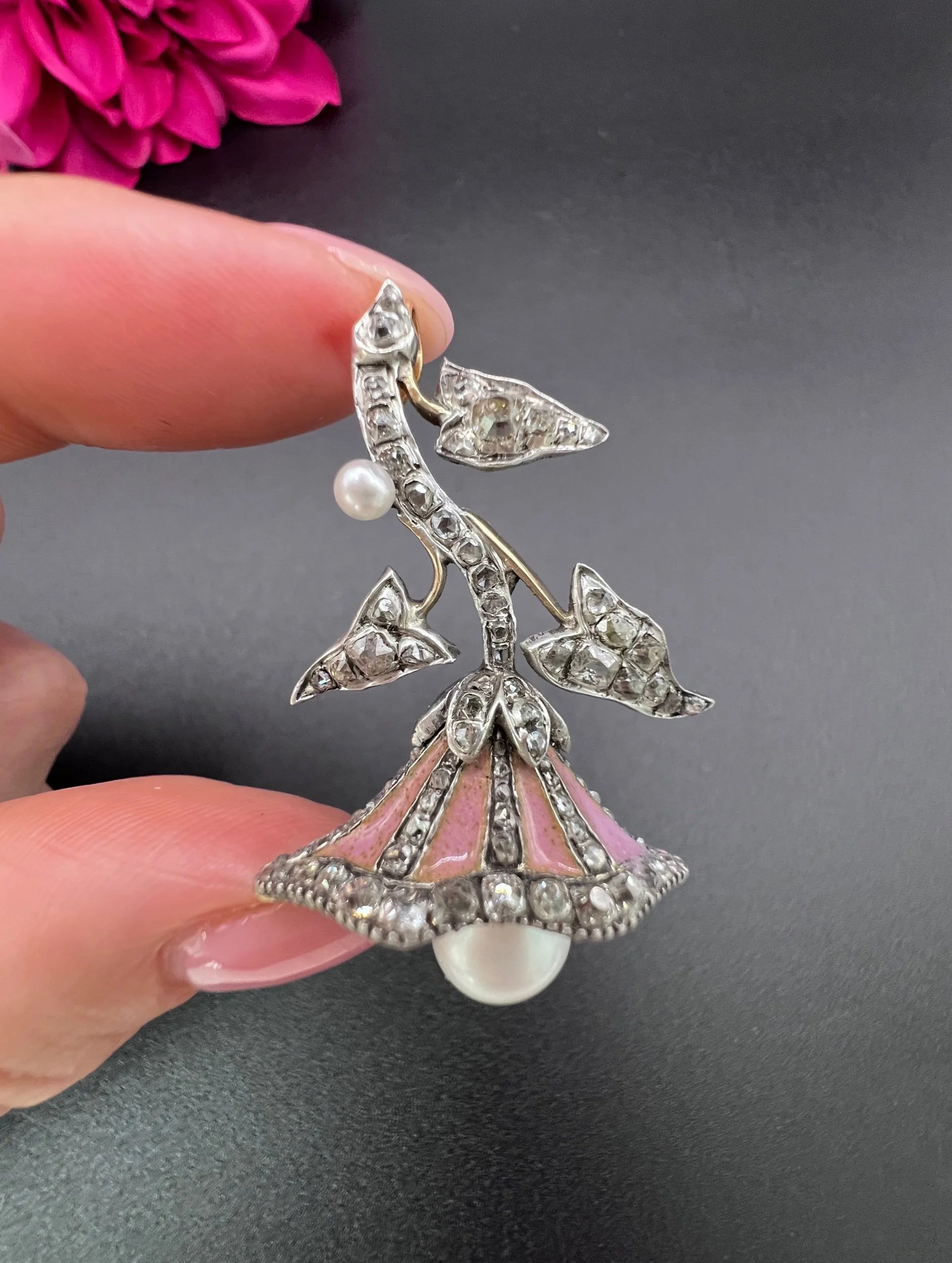 A pink enamel, diamond and pearl flower pendant in gold and silver, held in a hand