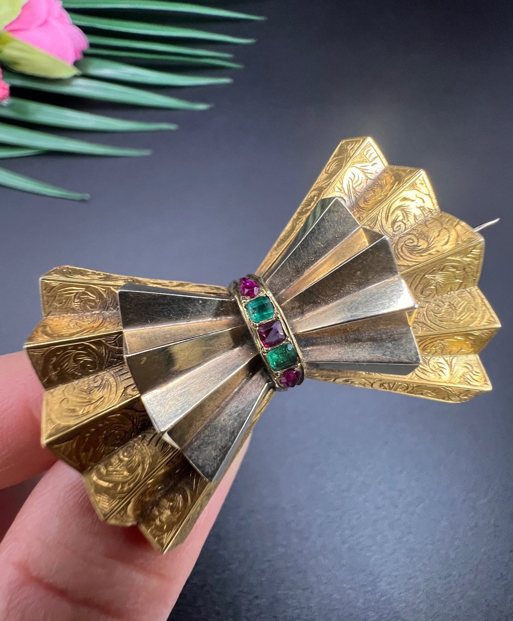 A yellow gold bow shaped brooch with rubies and emeralds, held in a hand