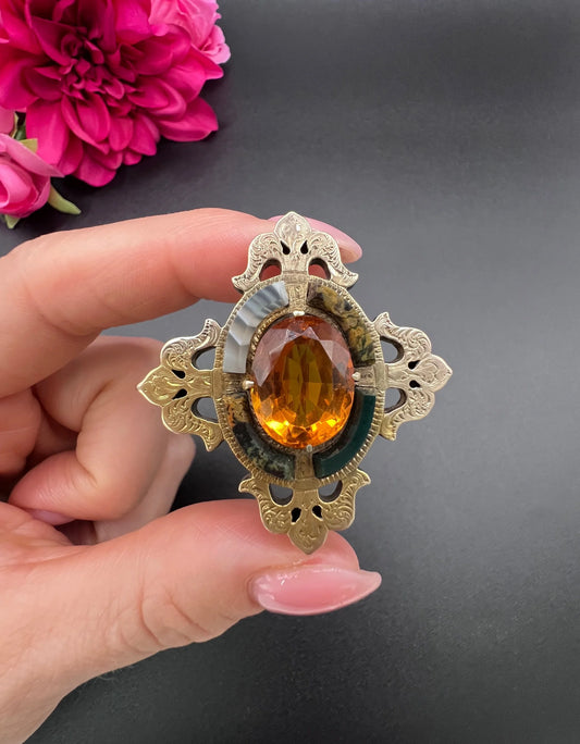 A yellow gold, oval citrine and agate ornate brooch, held in a hand