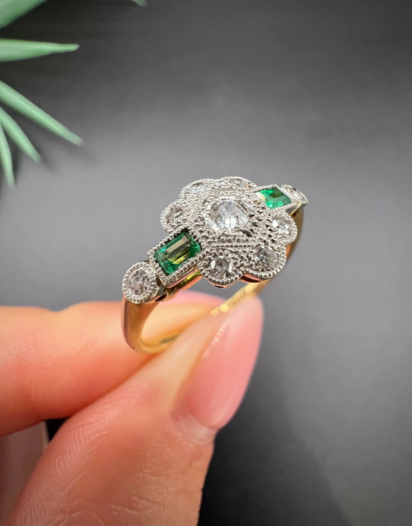 An emerald and diamond floral cluster ring in yellow gold and platinum, held in a hand