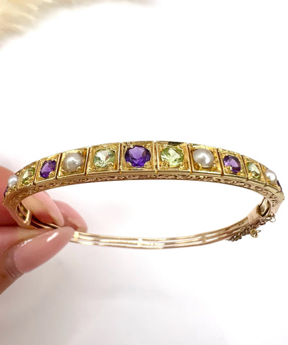A yellow gold peridot, amethyst and pearl bangle bracelet, held in a hand