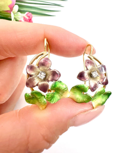 Gold and enamel diamond flower earrings held in a hand, against a white background
