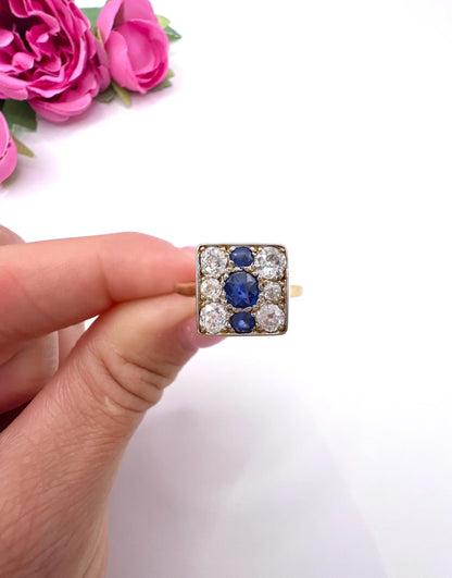 A square-shaped cluster ring with sapphires and diamonds in yellow gold and platinum, held in a hand