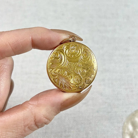 A circular yellow gold locket with engraving, held in a hand