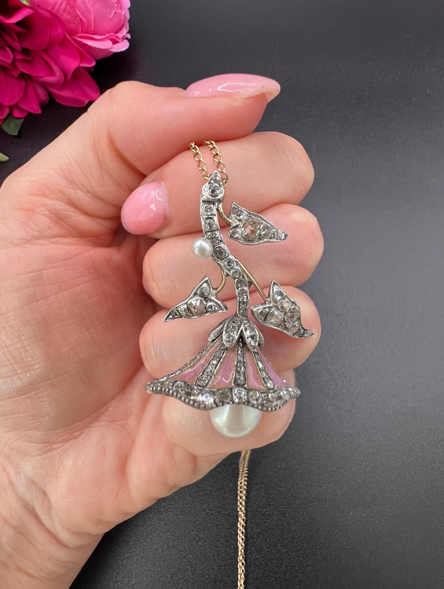 A pink enamel, diamond and pearl flower pendant in gold and silver, with a chain, held in a hand