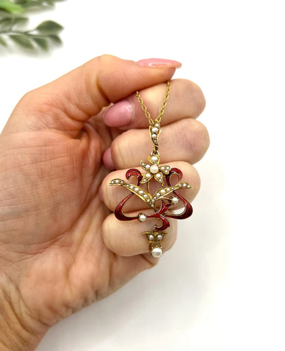A floral pendant in yellow gold with seed pearls and red enamel, with a chain, held in a hand