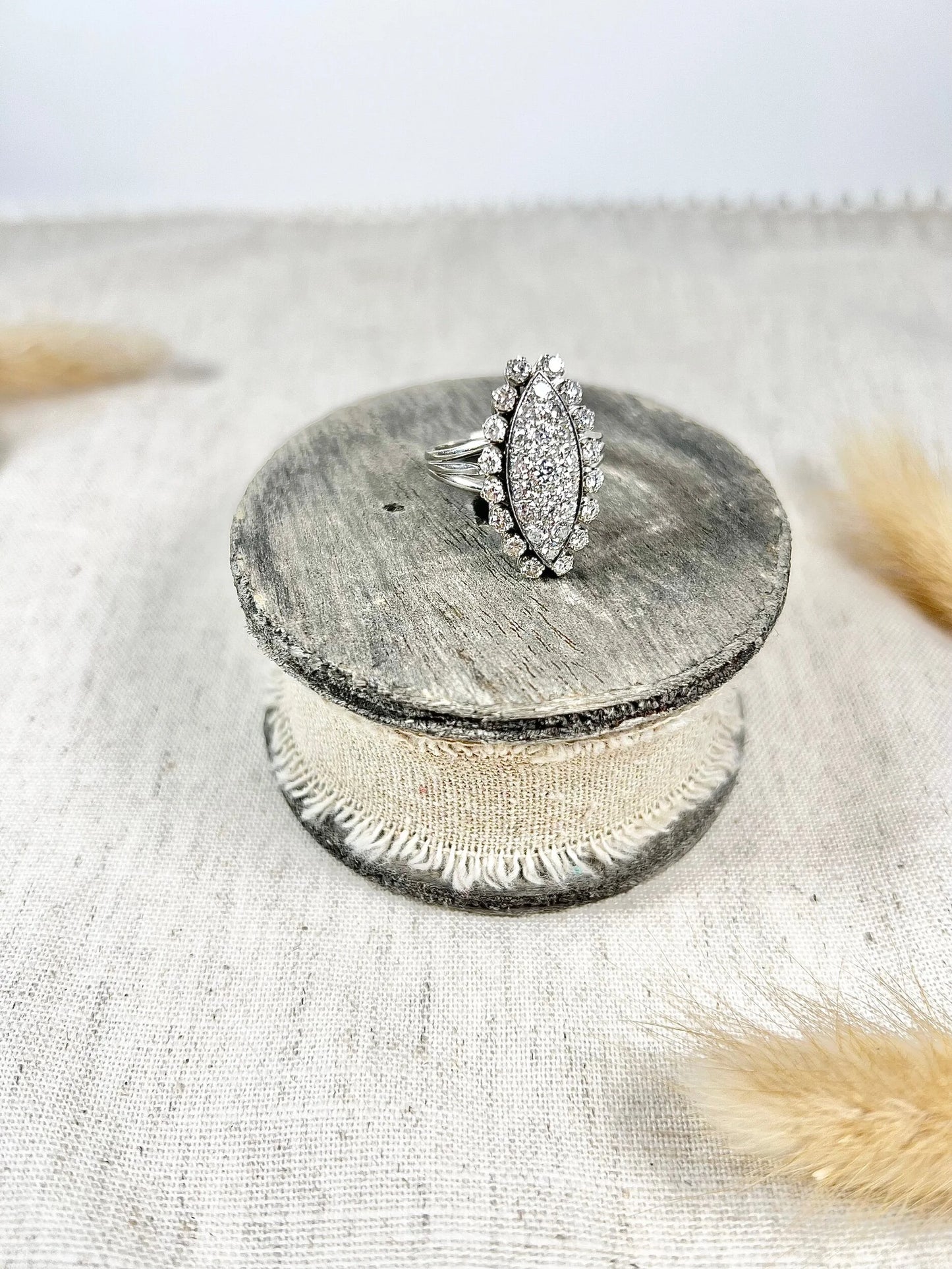 A navette shaped diamond cluster ring in white metal, sitting on top of a wooden and cloth object