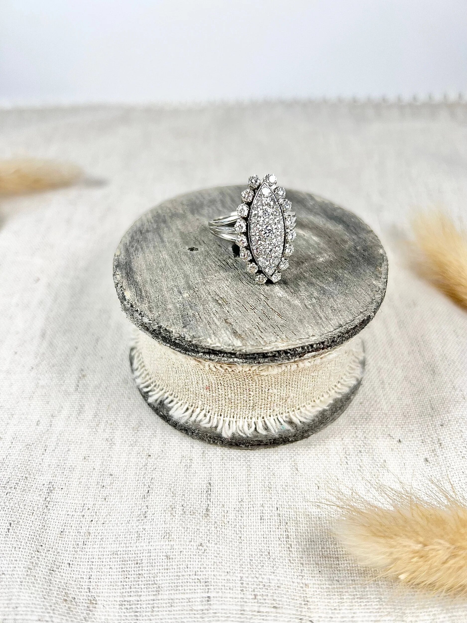A navette shaped diamond cluster ring in white metal, sitting on top of a wooden and cloth object