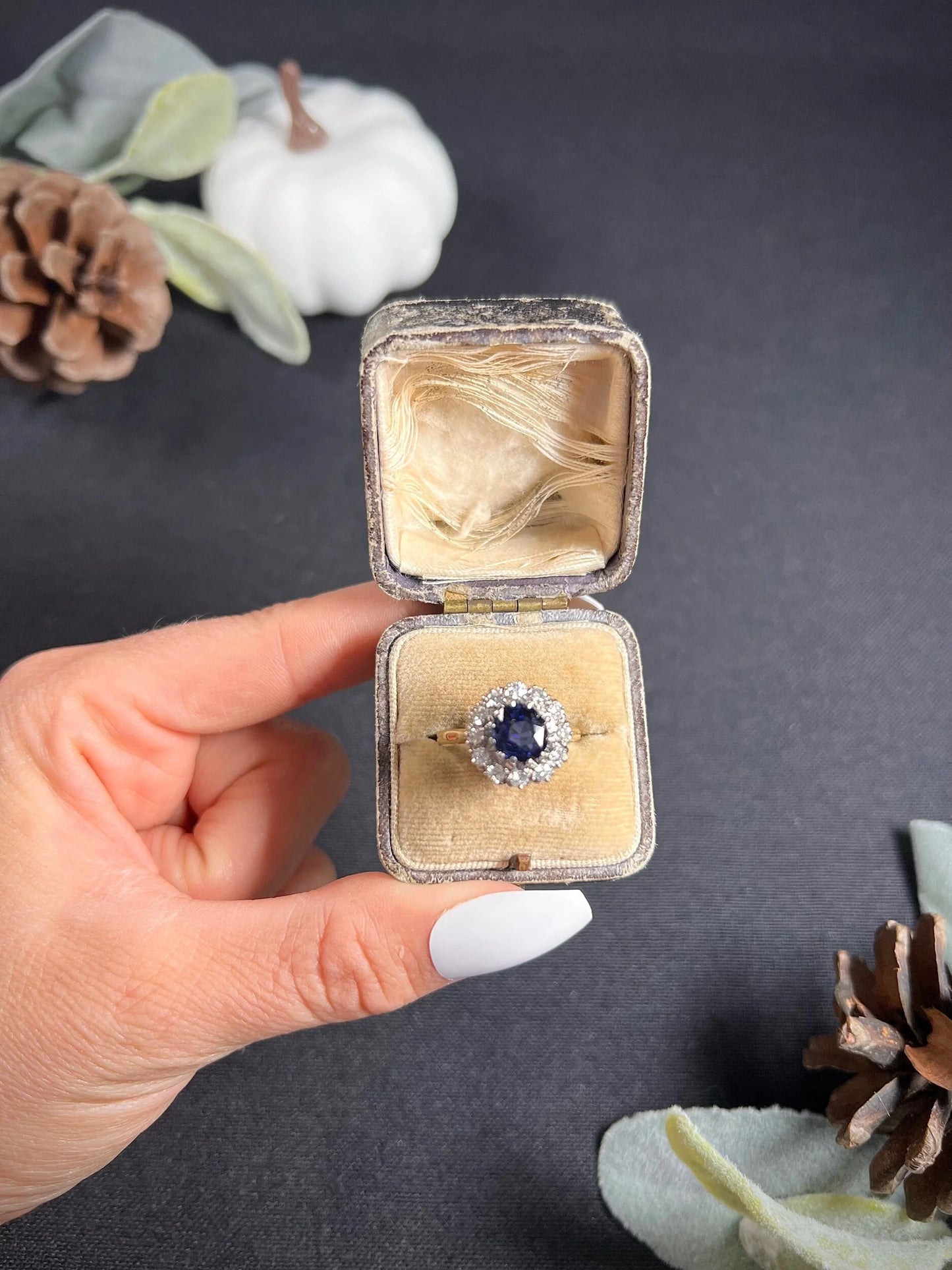A round sapphire and diamond cluster ring, in an antique ring box, held by a hand
