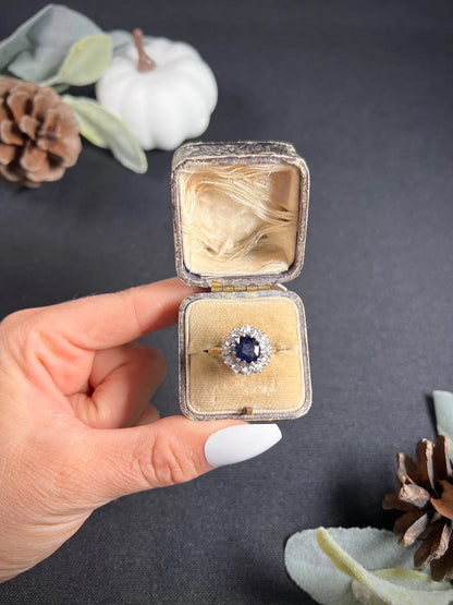 A round sapphire and diamond cluster ring, in an antique ring box, held by a hand
