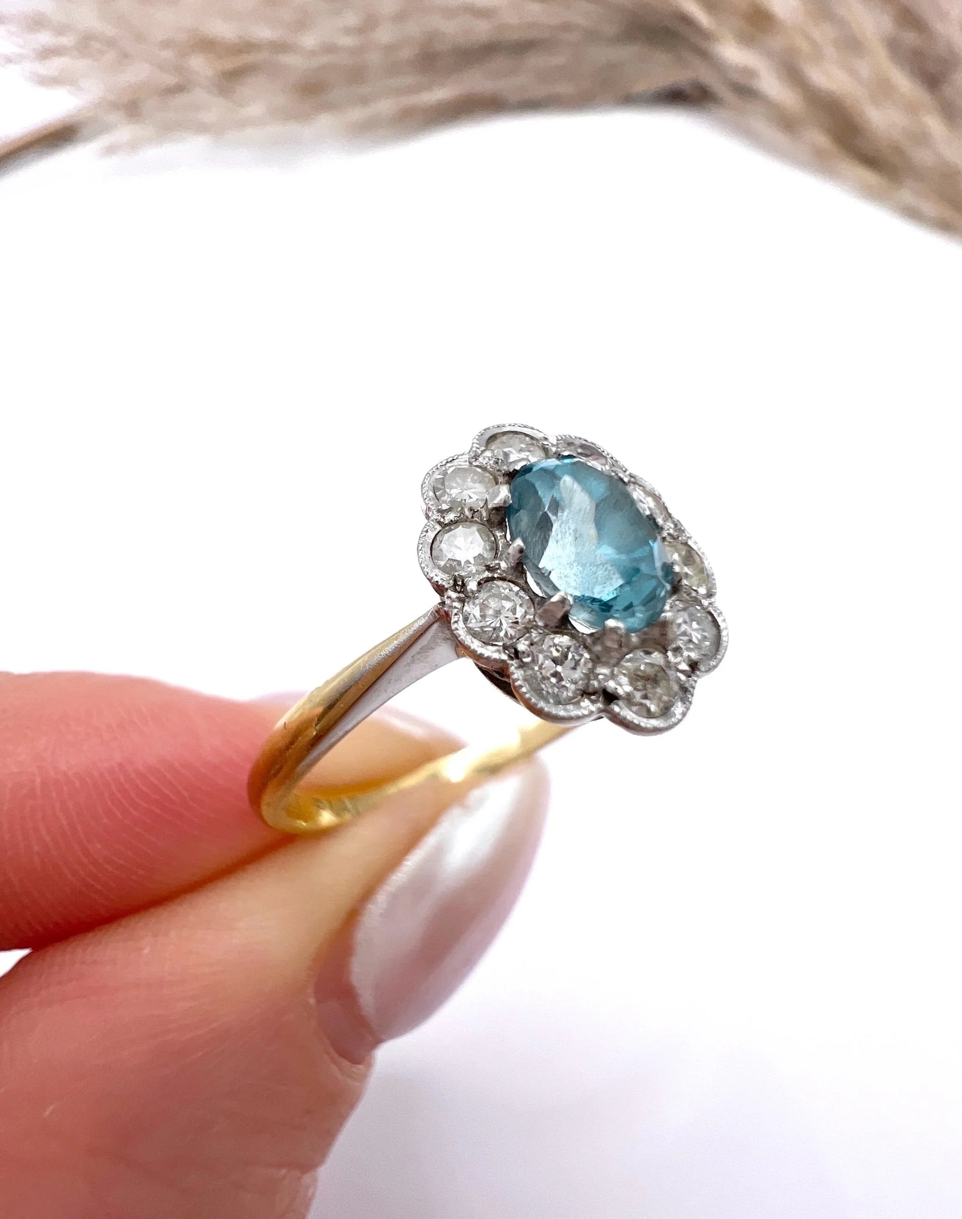 An oval cluster ring with a blue zircon centre surrounded by round diamonds, in yellow gold and platinum, held in a hand