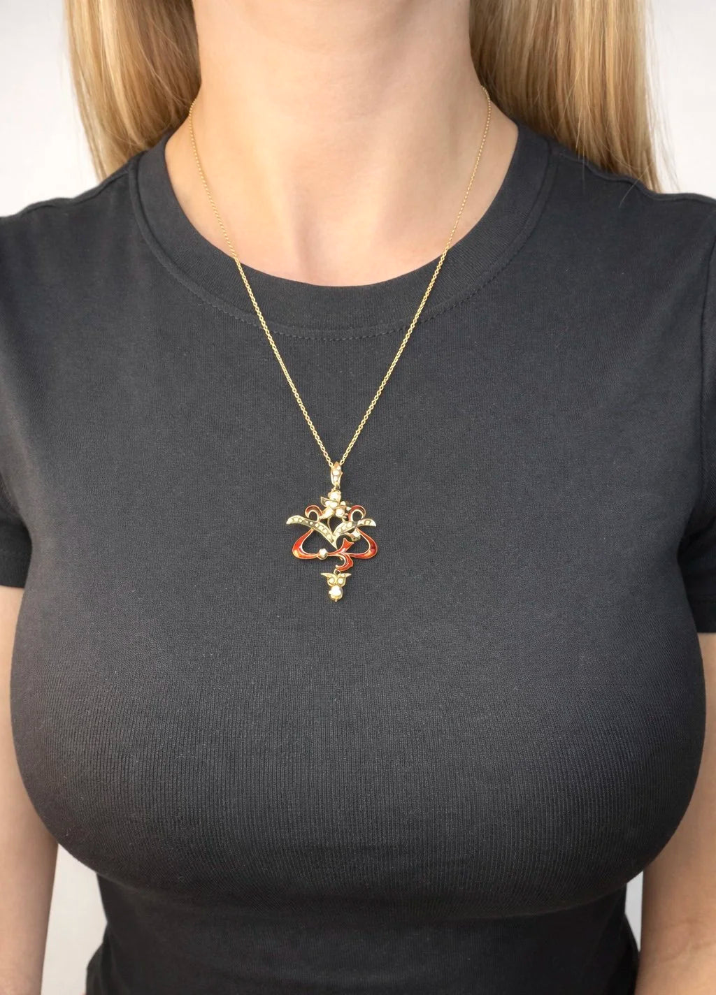 A floral pendant in yellow gold with seed pearls and red enamel, with a chain, modelled by a woman wearing a black top