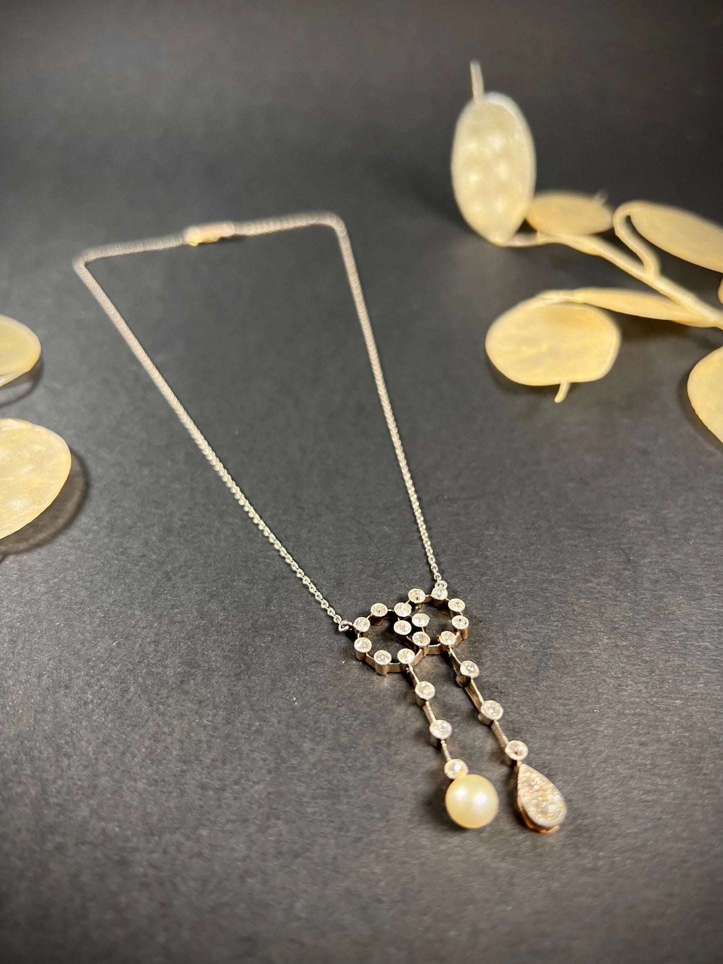 An antique diamond and pearl double drop pendant necklace, on a black background with dried leaves