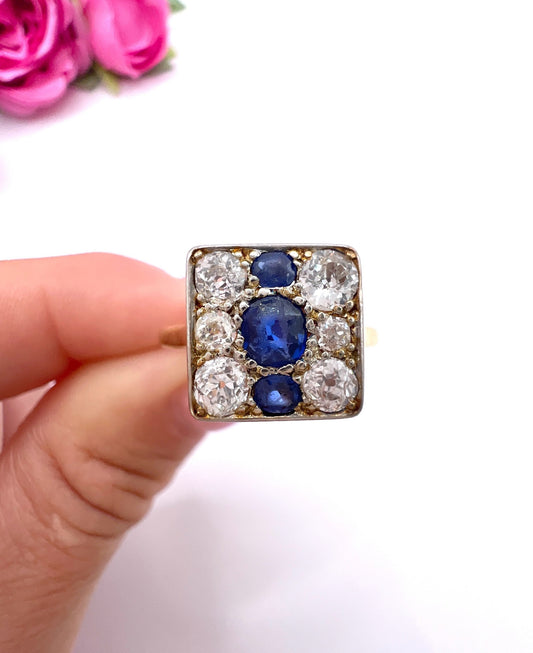 A square-shaped cluster ring with sapphires and diamonds in yellow gold and platinum, held in a hand