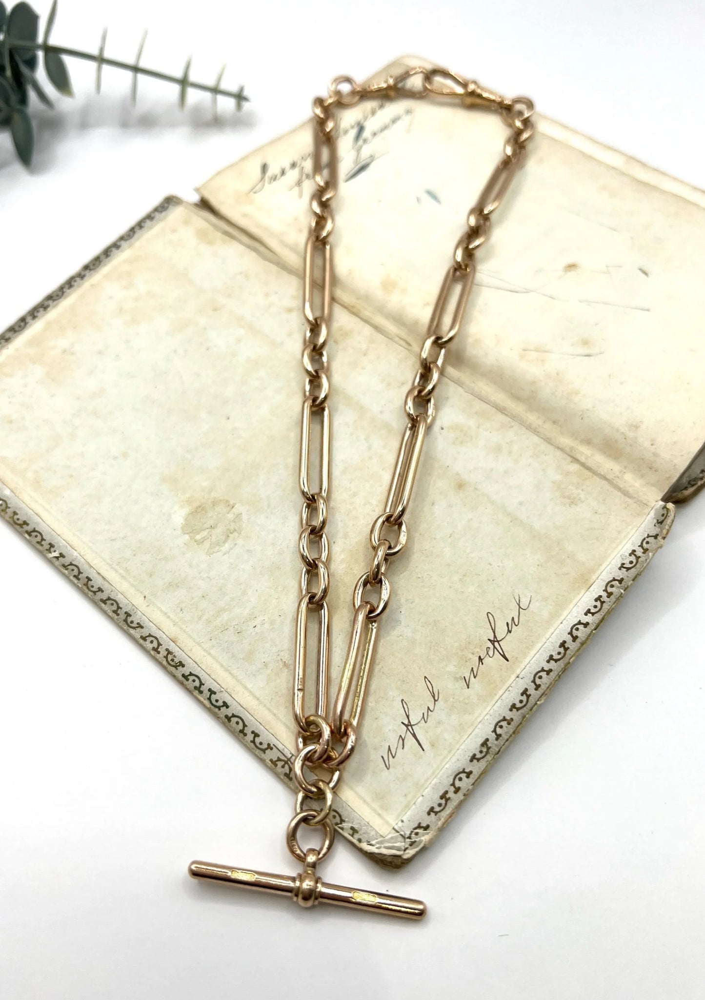 A rose gold trombone and fetter link Albert chain lying on top of an antique book, on a white background