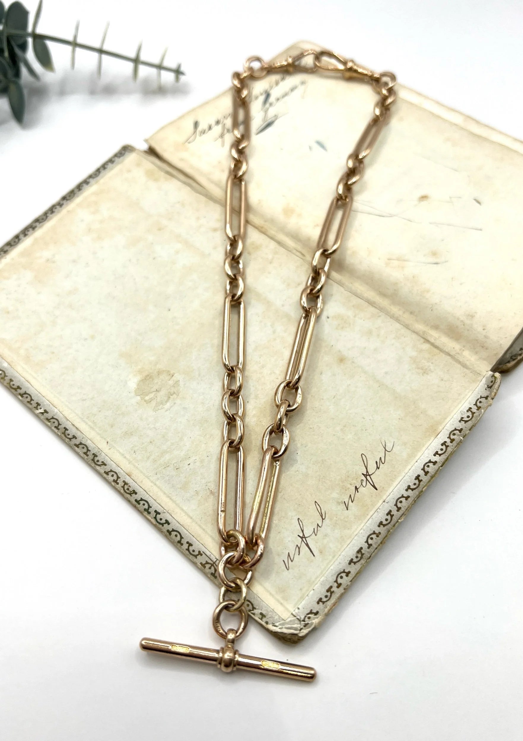 A rose gold trombone and fetter link Albert chain lying on top of an antique book, on a white background