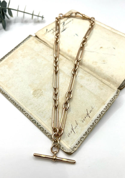 A rose gold trombone and fetter link Albert chain lying on top of an antique book, on a white background