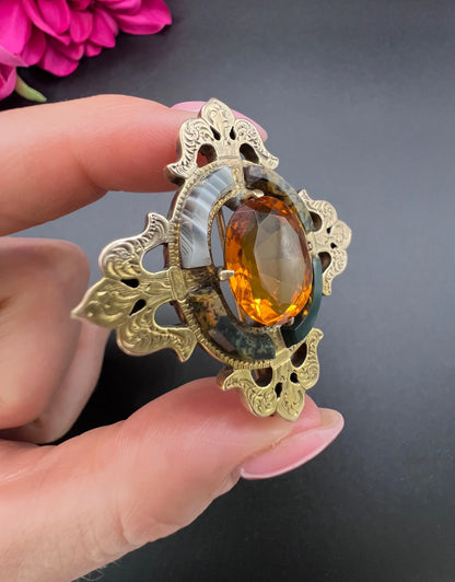 A yellow gold, oval citrine and agate ornate brooch, held in a hand