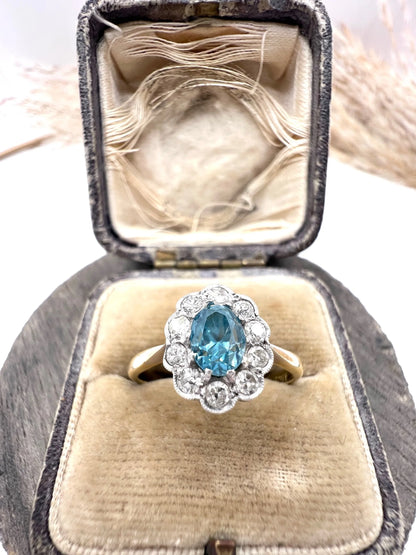 An oval cluster ring with a blue zircon centre surrounded by round diamonds, in yellow gold and platinum, in an antique ring box