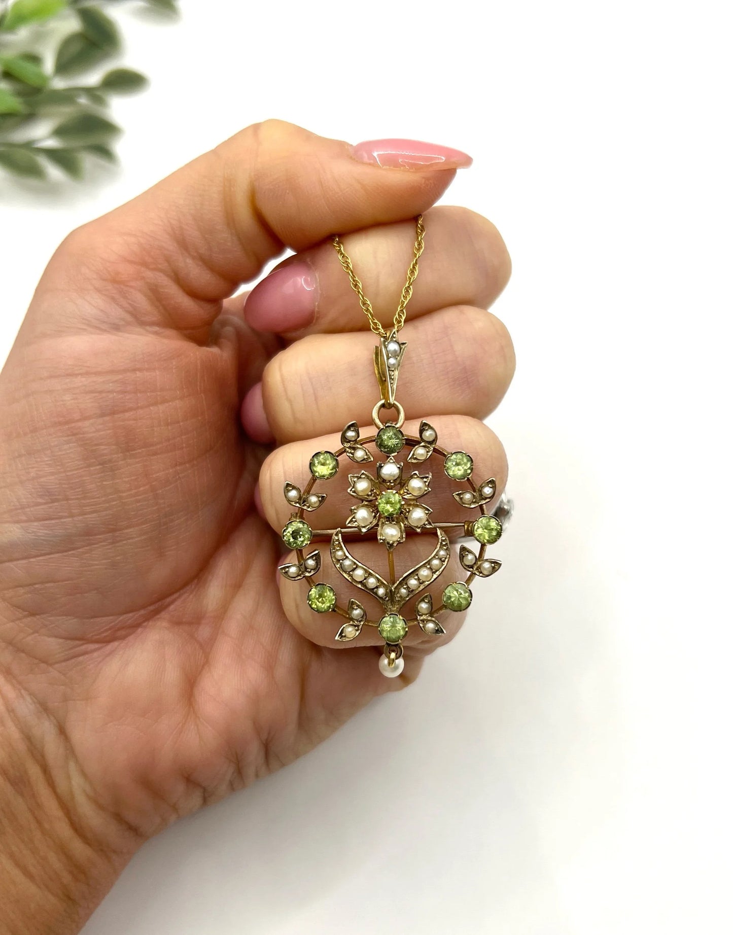 A peridot and pearl floral circle brooch/pendant in yellow gold, with a chain, held in a hand
