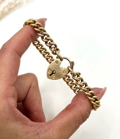 A gold curb link bracelet with heart shaped padlock, held in a hand