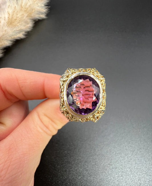 A yellow and white gold ornate solitaire ring with an oval amethyst, held in a hand