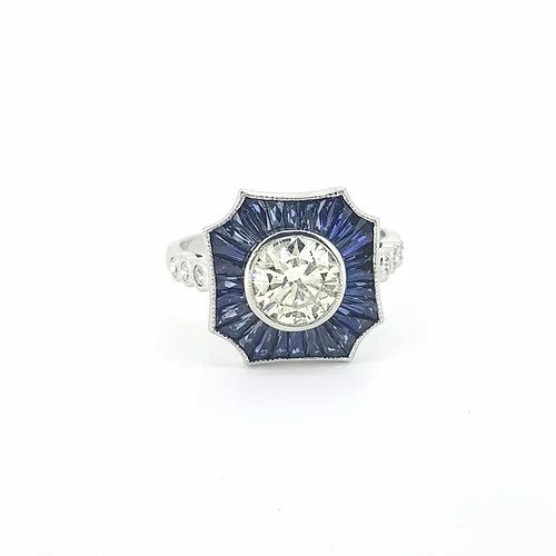 Contemporary Diamond and Sapphire Cluster Target Ring; central 1.01 carat round brilliant-cut diamond surrounded by calibre cut sapphires, three diamonds to each shoulder, in 18ct white gold.