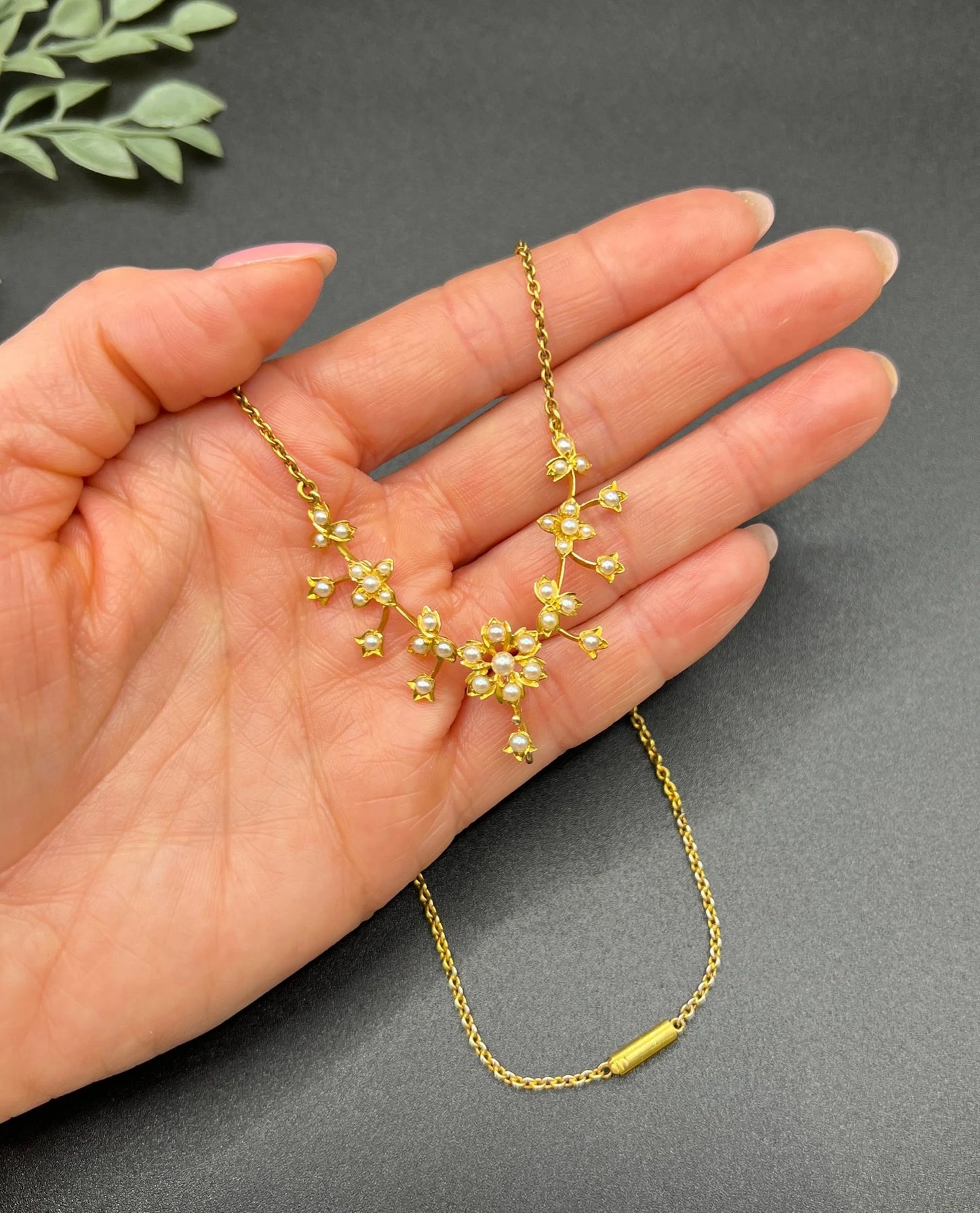 A yellow gold floral pendant necklace with seed pearls, held in a hand
