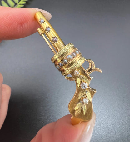 A yellow gold novelty gun brooch with diamonds and seed pearls, held in a hand