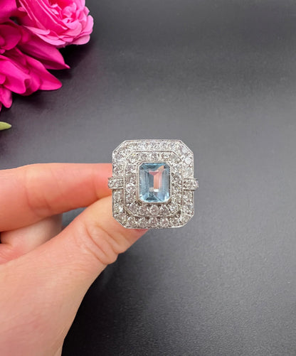 An octagonal shaped cluster ring with an aquamarine centre surrounded by two rows of diamonds, in platinum, held in a hand