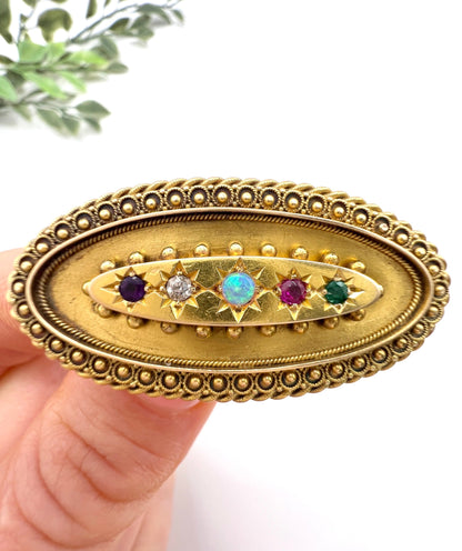 An oval yellow gold multi-gemstone acrostic brooch, held in a hand