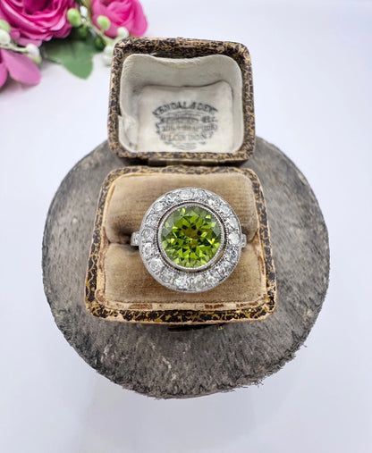 A white gold circular target cluster ring with a peridot centre surrounded by diamonds, in a ring box
