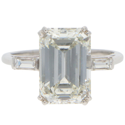 A large emerald cut diamond ring with baguette cut diamond shoulders in white metal, on a white background