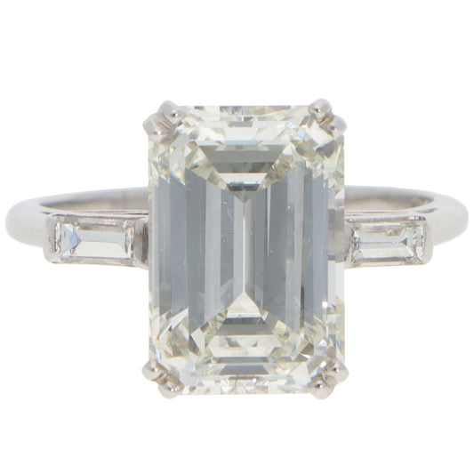 A large emerald cut diamond ring with baguette cut diamond shoulders in white metal, on a white background